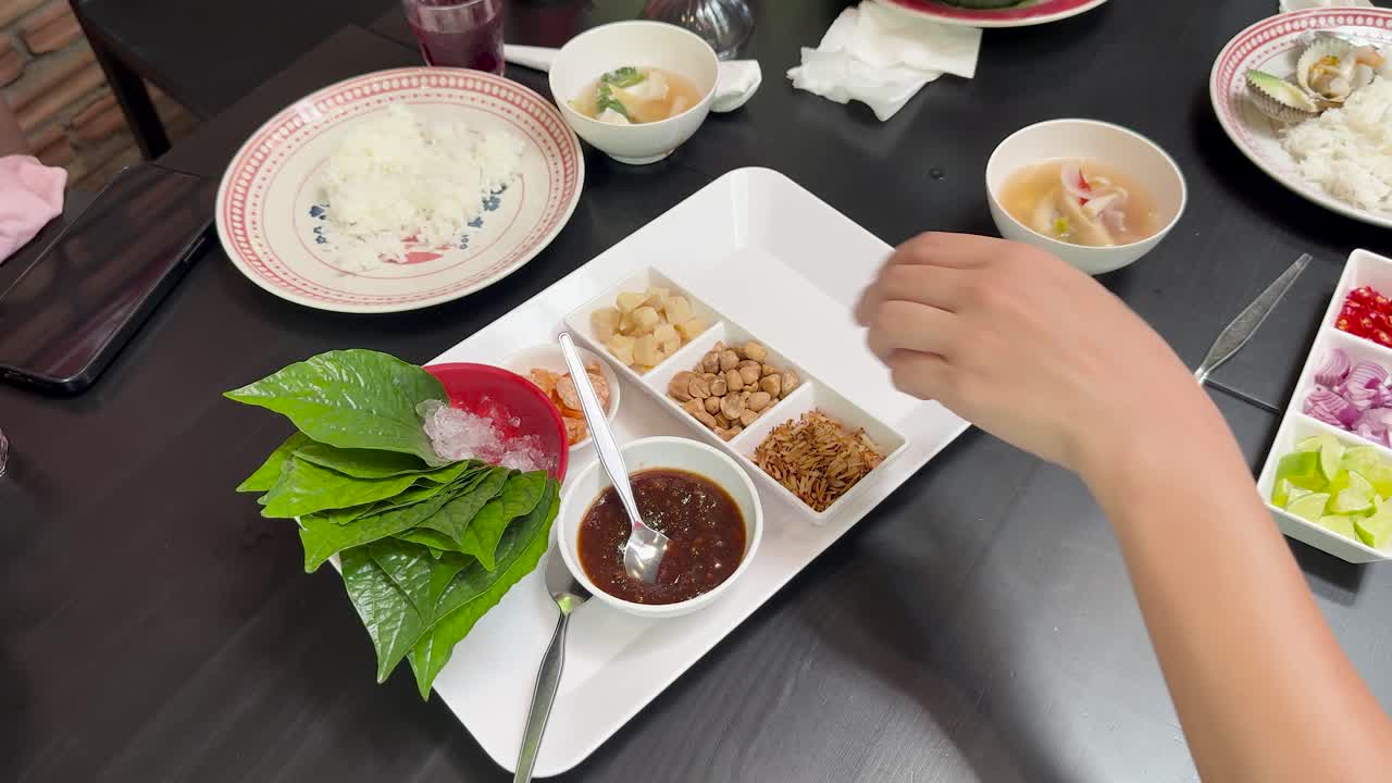 Two people assemble Miang Kham, a traditional Thai snack, using fresh leaves, assorted fillings, and sauce under bright, even lighting at a modern dining table