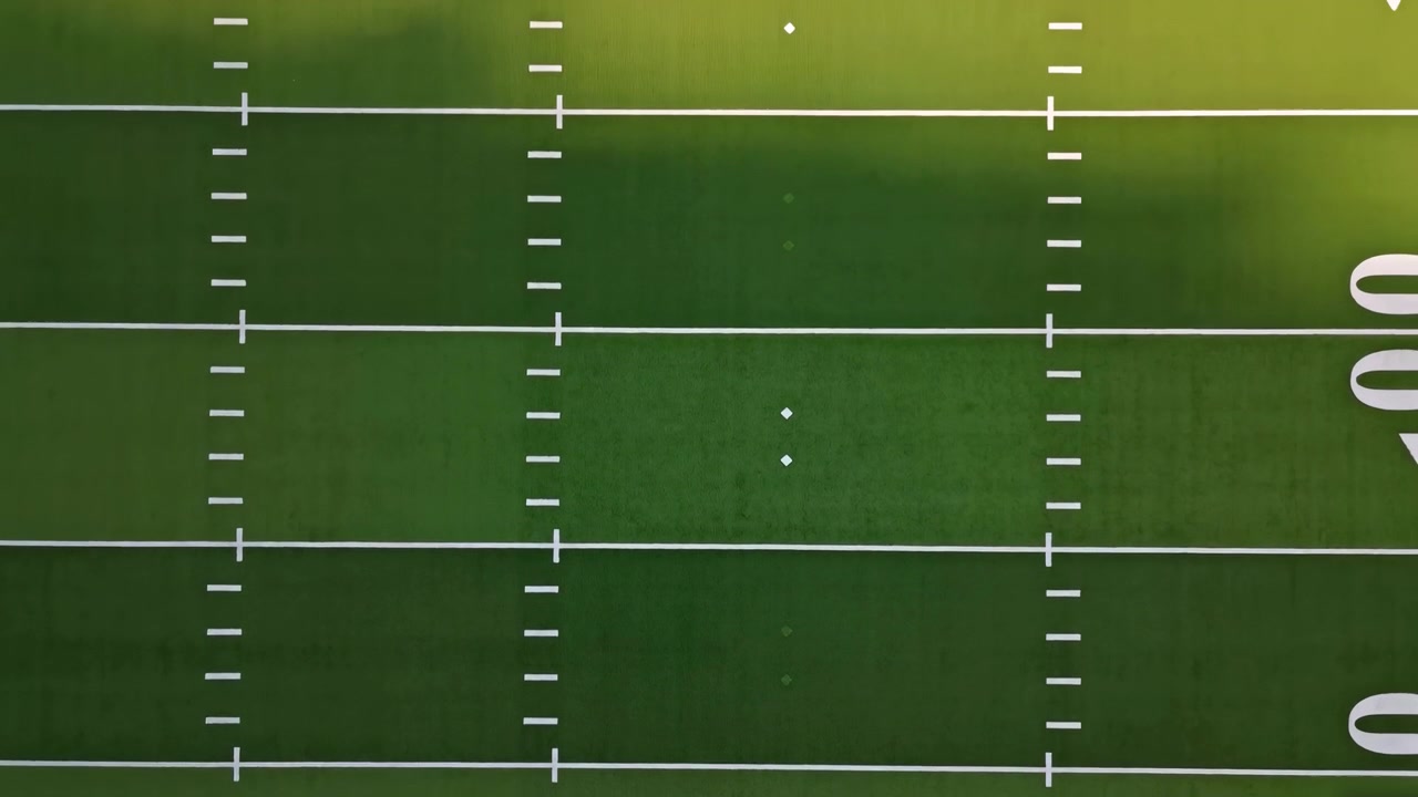 Una vista aerea diretta rivela il centrocampo vuoto di un campo da football americano con erba verde, doppie linee delle yard, tacche bianche e una simmetria nitida per uno sfondo sportivo pulito e per uso televisivo.