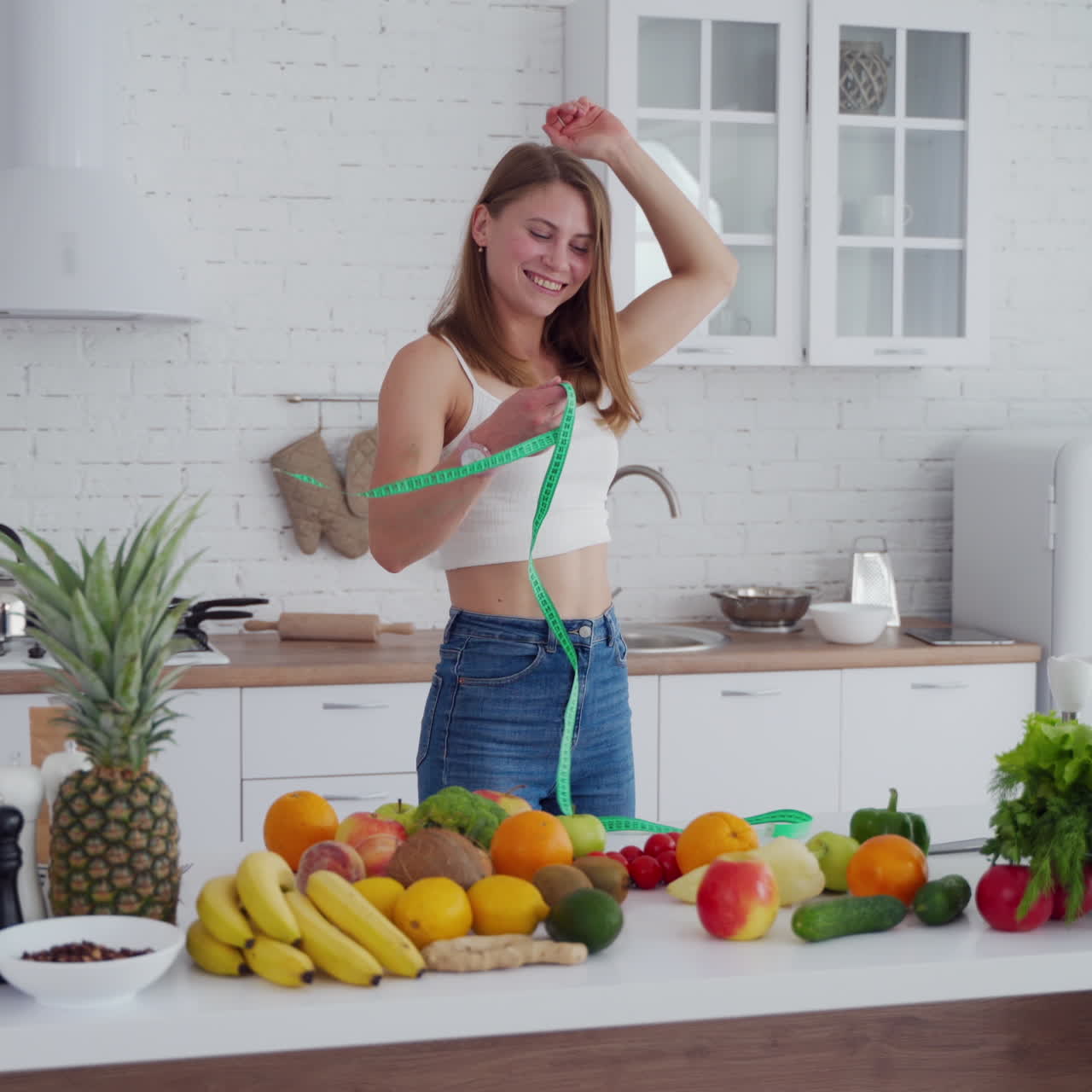 Slim woman measures her waist in the kitchen. Happy girl in jeans standing near the table full of fresh fruit and vegetables at home. Young lady loses weight.