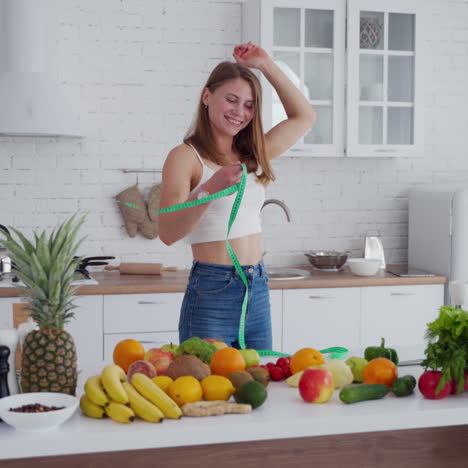 slim woman measures her waist in the kitchen. happy girl in jeans standing near the table full of fresh fruit and vegetables at home. young lady loses weight.
