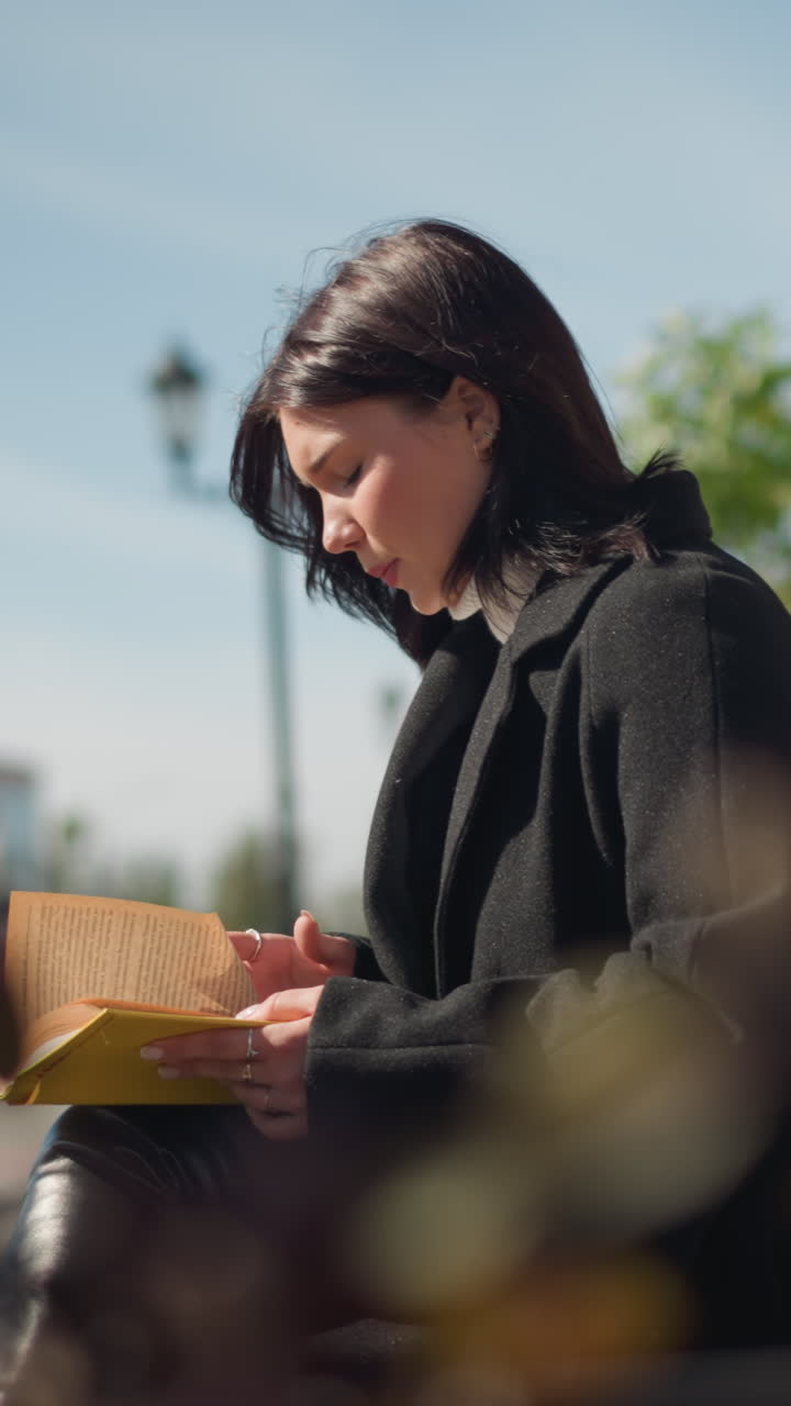 joven con abrigo negro sentada al aire libre en un banco leyendo un libro con enfoque, edificios urbanos y árboles en el fondo soleado, la luz del sol suave iluminando su expresión pensativa