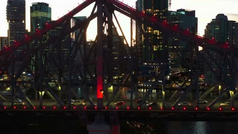 story bridge adventure climb, tiny people climbing on iconic cantilever bridge with busy traffic crossing the river at dusk and illuminated downtown cityscape of brisbane city, cinematic aerial shot