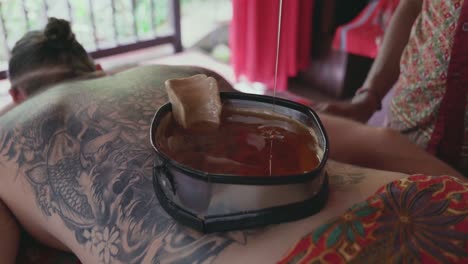 spa therapist pours a steady stream of warm oil into a wax-lined container on the back of a male patient enjoying a kati vasti ayurvedic treatment