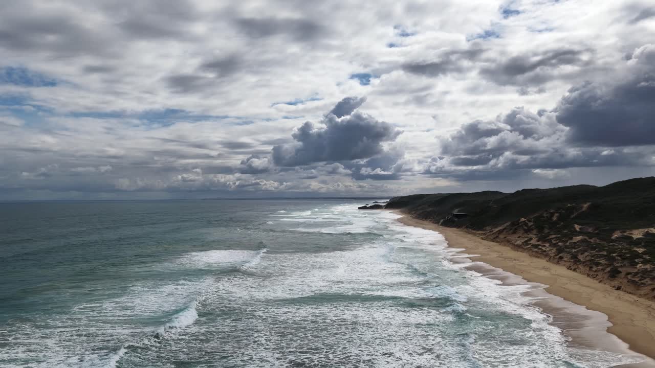 Drone captures dramatic waves, sandy shoreline, and moody clouds along Portsea coast, natural daylight