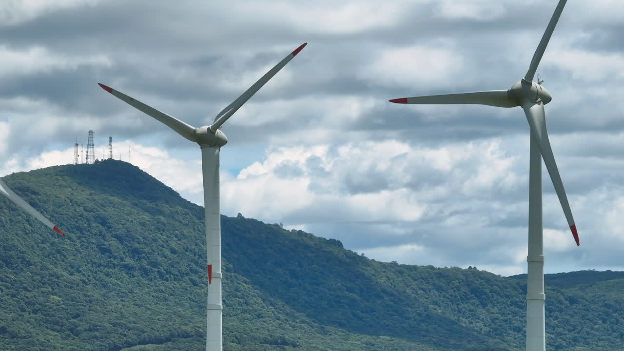 Professional static aerial shot of wind turbines spinning against a lush green hill with communication antennas. Concept of clean power and modern infrastructure. Mavic 3 4K 10-bit D-Log video