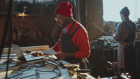 african american blacksmith using laptop and taking notes in workshop