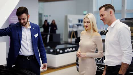 salesman showing car engine under open hood to couple in auto showroom