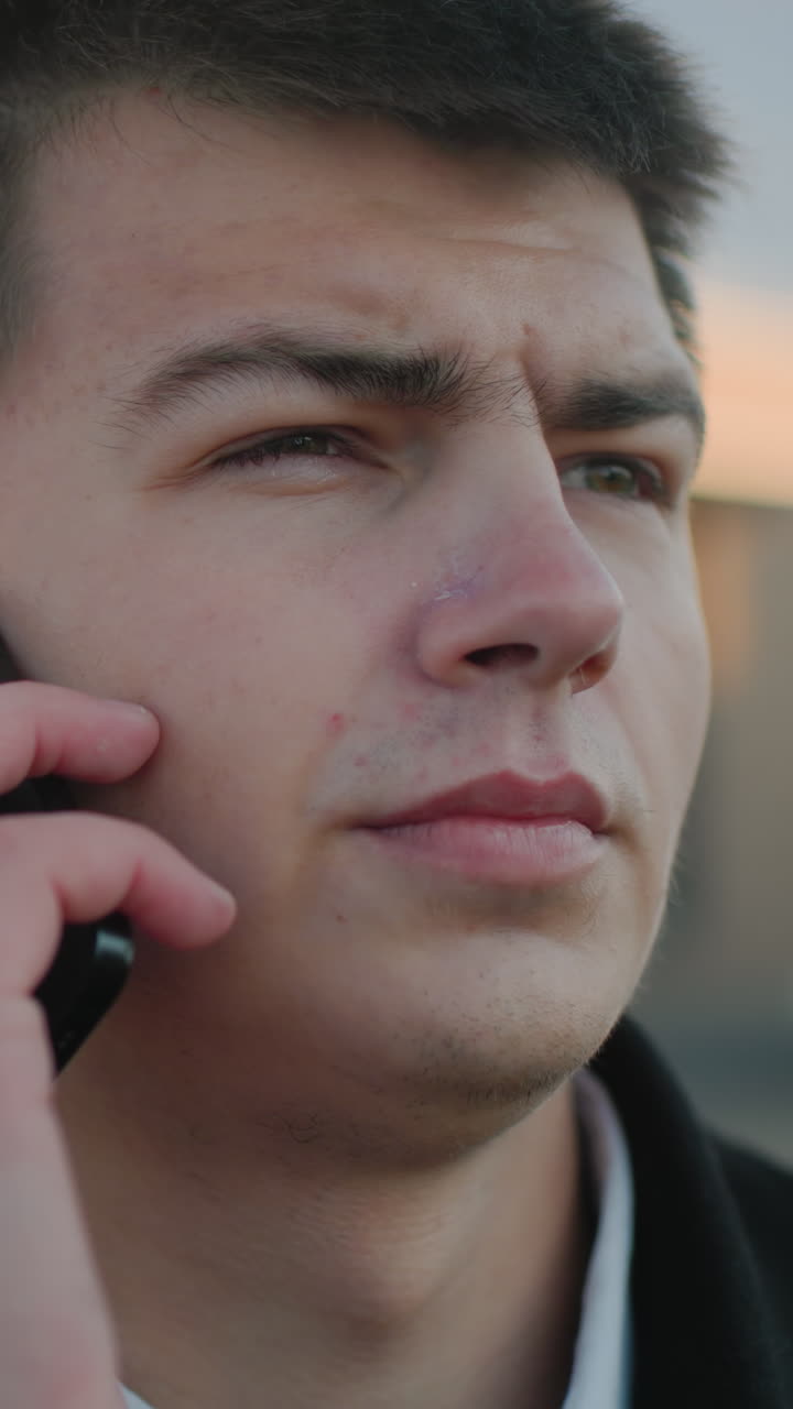 흑색 코트와 색 셔츠를 입고 부드러운 조명과 흐릿한 도시 배경으로 전화 통화에 종사하는 남자의 클로즈업, 표정은 야외 도시 환경에서 전문성을 반영합니다.