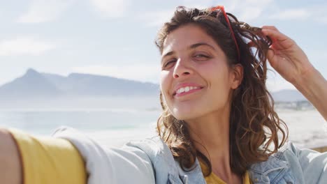 feliz mujer de raza mixta haciendo un video selfie saludando y sonriendo en el soleado paseo marítimo junto al mar