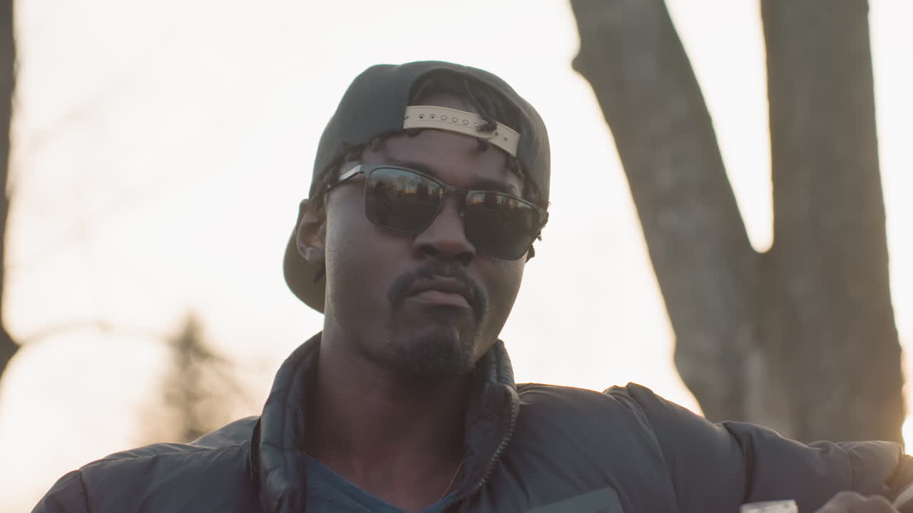 Young man wearing sunglasses and backward cap drinks orange juice from plastic bottle in park during golden hour, relaxed expression, casual jacket, warm backlight
