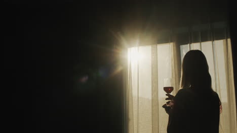 A-woman-with-a-glass-of-wine-stands-at-the-window-at-sunset