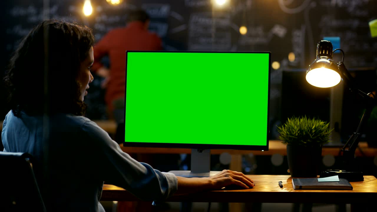 Beautiful Female Office Employee at Her Desktop Works on a Mock-up Green Screen Personal Computer. Over the Shoulder Footage. Her Colleague works in the Background. Creative Office Evening.