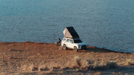 white car with roofnest tent and man sitting next to lake, aerial