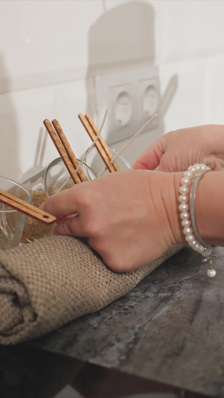 Primo piano delle mani di una donna che sistema supporti in legno per stoppini e stampi in vetro per candele su un bancone. La scena include tessuto di iuta arrotolato, bicchieri trasparenti con materiale granulare all'interno, illuminazione soffusa.