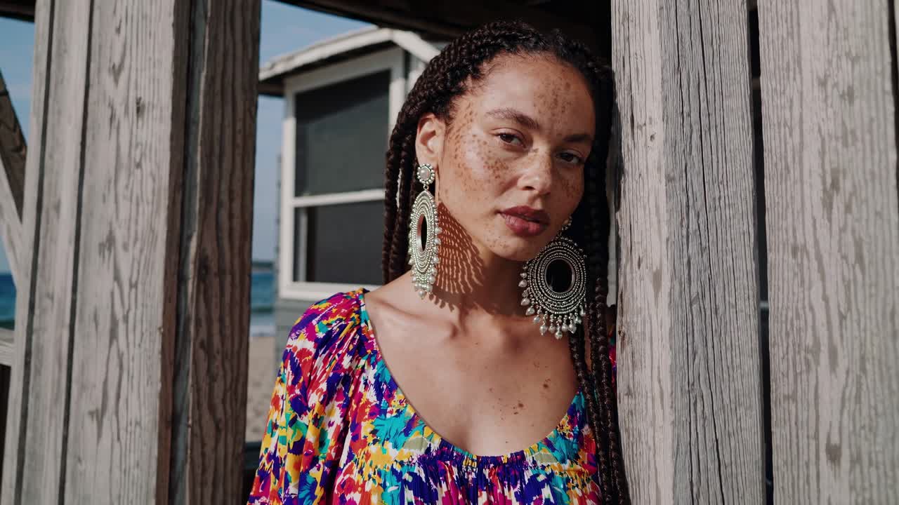 Capturing a confident woman with braided hair and bold earrings, embodying effortless beach style against a rustic wooden backdrop. The colorful dress enhances her vibrant summer look