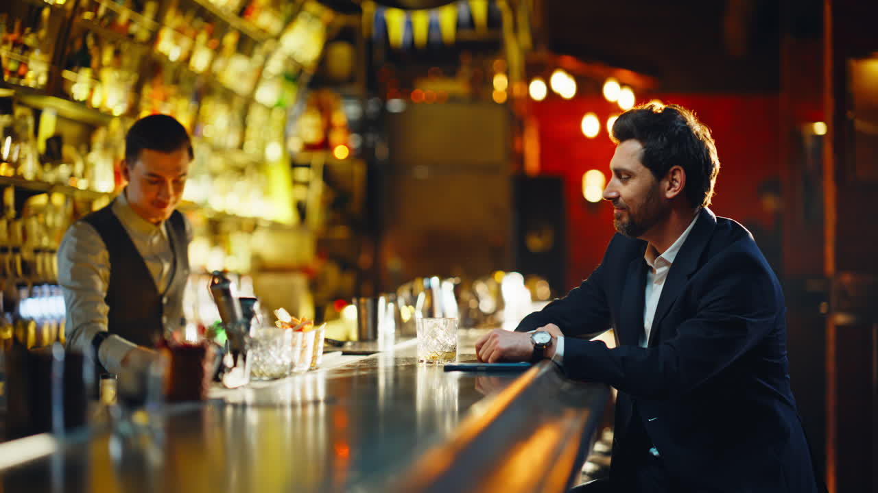 Primo piano di un cliente in un pub che gusta un bicchiere di bourbon di sera. Un signore sorridente sta pagando