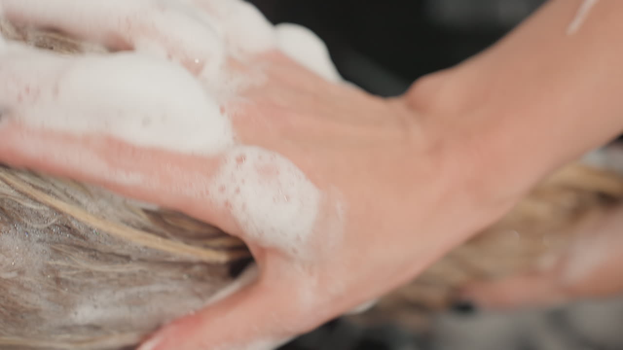 Parrucchiere che lava capelli biondi con shampoo schiumoso ed esegue un delicato massaggio al cuoio capelluto, primo piano delle dita che massaggiano le radici, schiuma cremosa sui capelli bagnati, lavabo pronto per l'asciugamano, illuminazione rilassante da spa e atmosfera distensiva