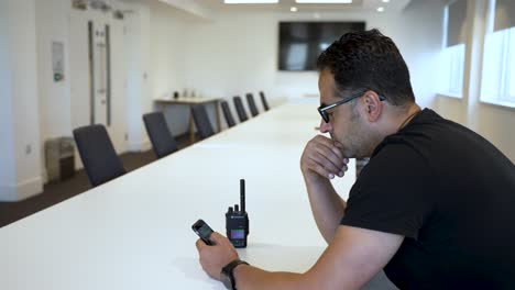 se ve a un hombre con una camisa negra sentado en una oficina moderna, concentrado en el uso de un teléfono inteligente