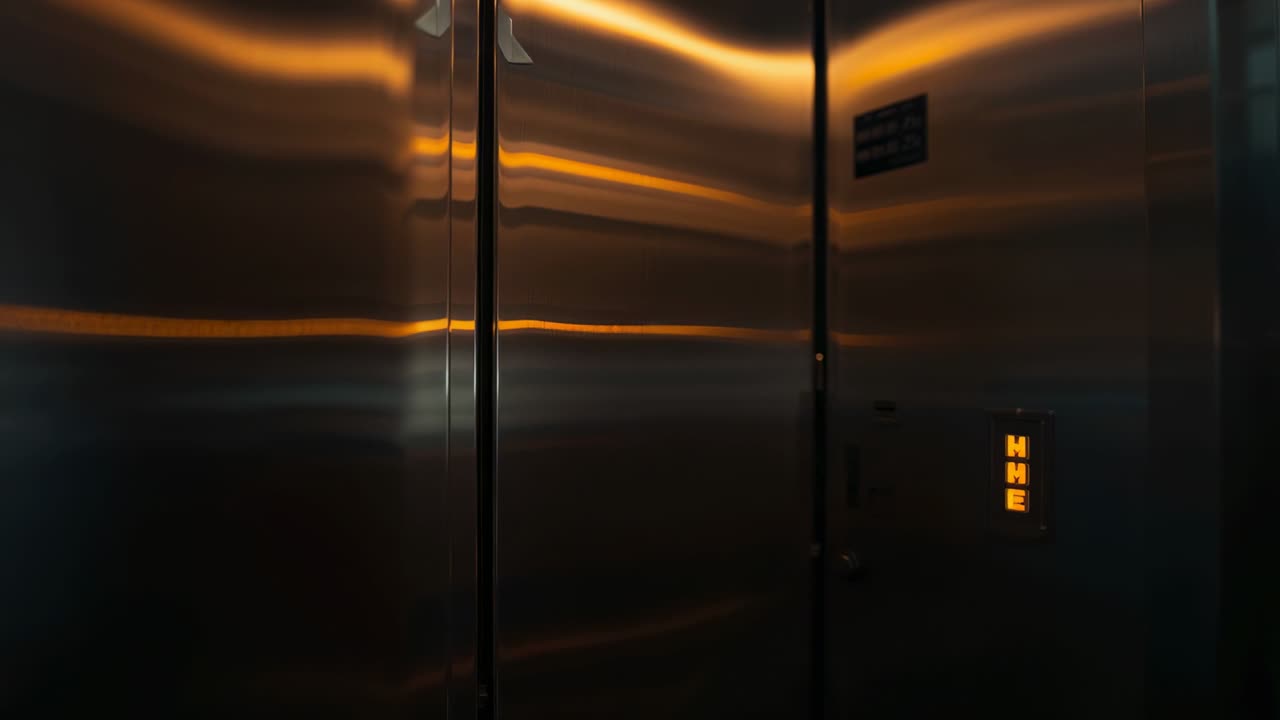 Menampilkan pintu lift logam memantulkan garis-garis amber di lobi, dengan indikator oranye, plakat, dan selotip.