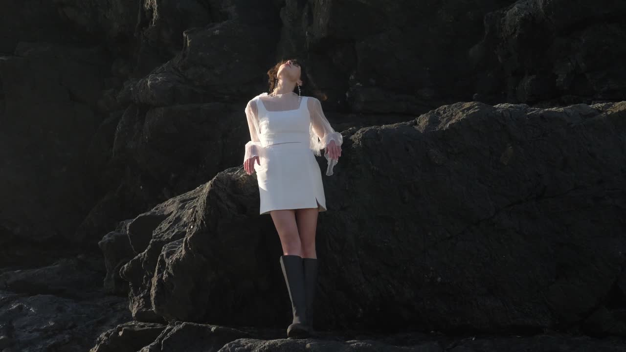 Stunning Woman In White Square-neck Dress With Sheer Sleeves Gracefully Strolls On Beach Rocks With Elegance And Poise