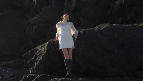 mujer impresionante en vestido blanco de cuello cuadrado con mangas transparentes graciosamente pasea por las rocas de la playa con elegancia y equilibrio