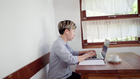 mujer adulta de cabello corto se sienta y toma notas para una reunión en línea con una computadora portátil en un café durante el día