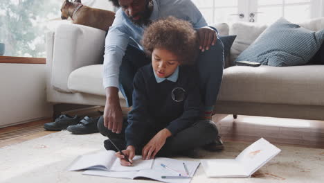 Low-angle-view-of-pre-teen-mixed-race-boy-in-school-uniform-sitting-on-the-floor-at-home-doing-his-homework,-with-his-dad-sitting-behind-him-on-the-sofa-helping,-beside-their-pet-dog,-close-up