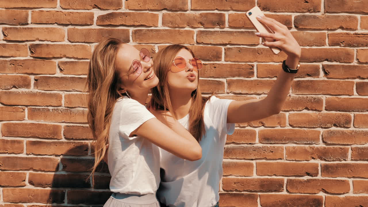 Dos jóvenes felices haciéndose un selfie frente a una pared de ladrillo
