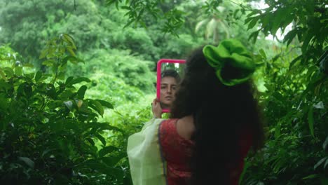 a woman in a green sari looks at herself in a pink mirror, surrounded by tropical vegetation