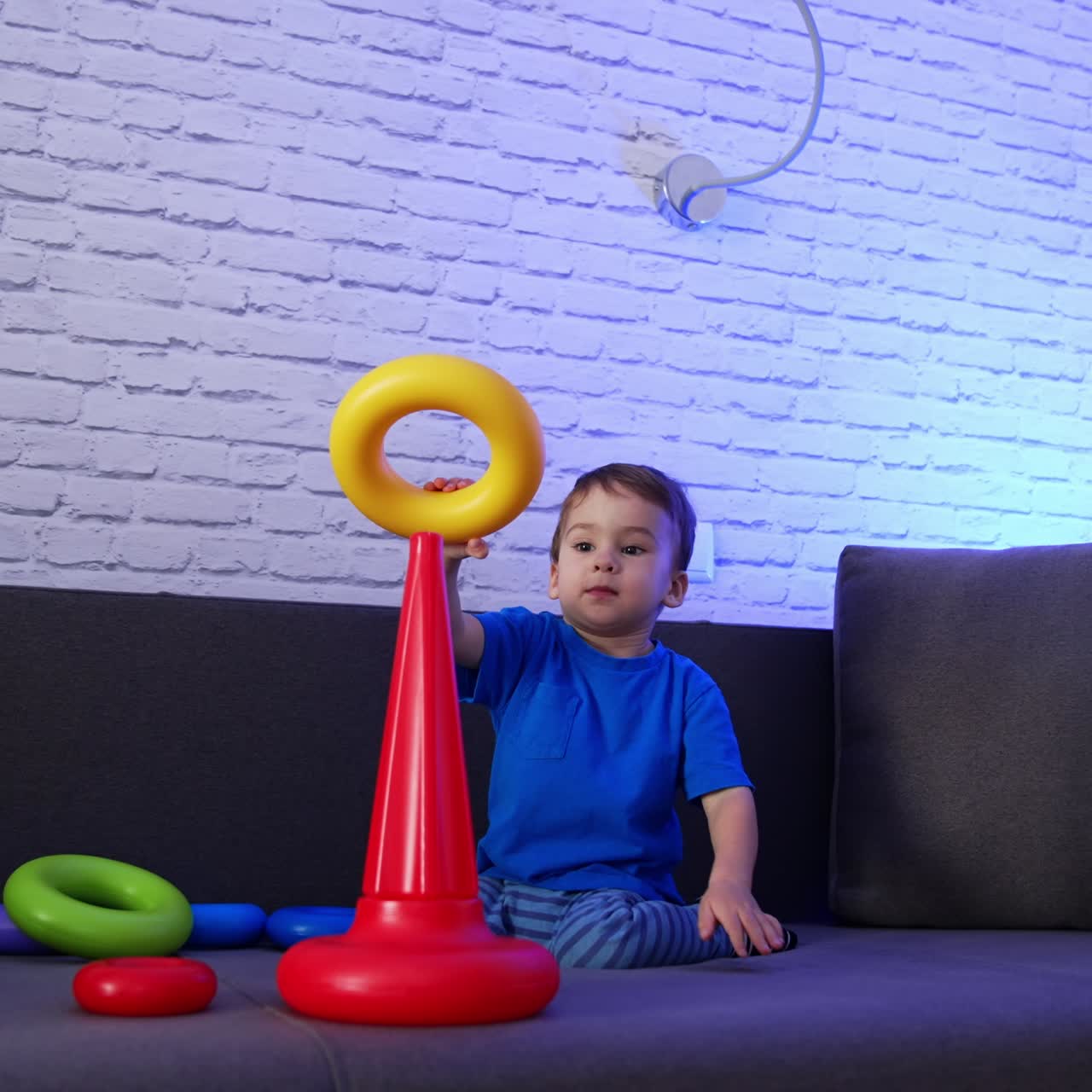 Cute dark-haired toddler assembling pyramid on the sofa. Baby boy plays with toy at home