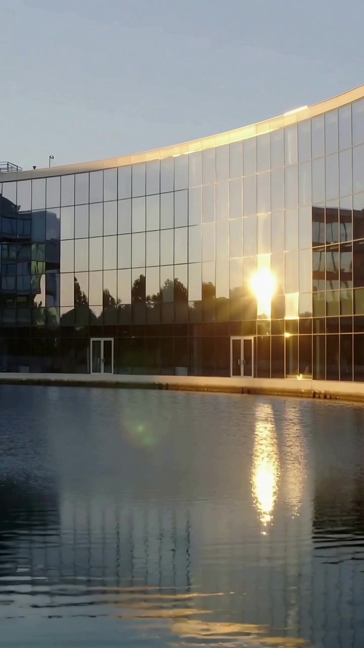 Vídeo vertical: Fachada de vidro cintilante reagindo ao brilho do sol, estendendo a reflexão na piscina da praça.