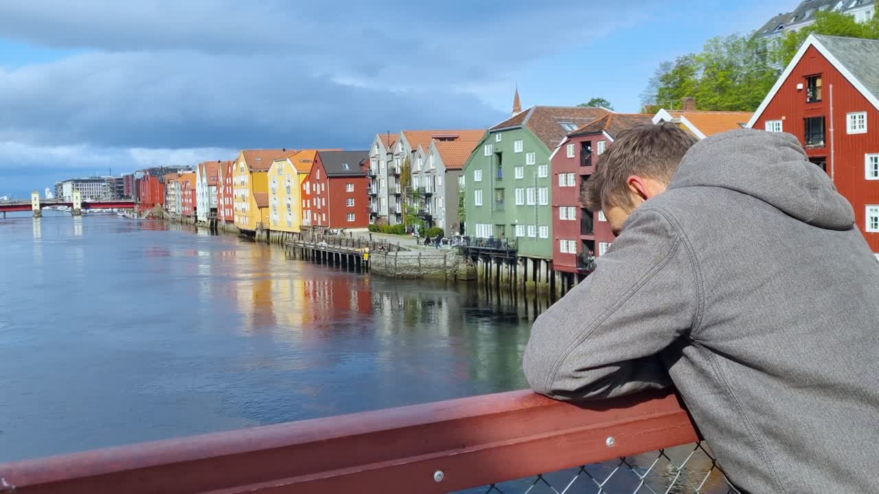 A man calmly enjoys the view of Trondheim’s colorful riverside stilt houses, their reflections shimmering in the still water on a sunny spring day in Norway