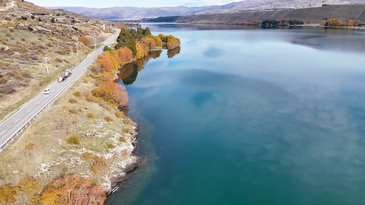 Drone flyover captures serene Lake Dunstan with autumn foliage and a winding road in Cromwell, New Zealand