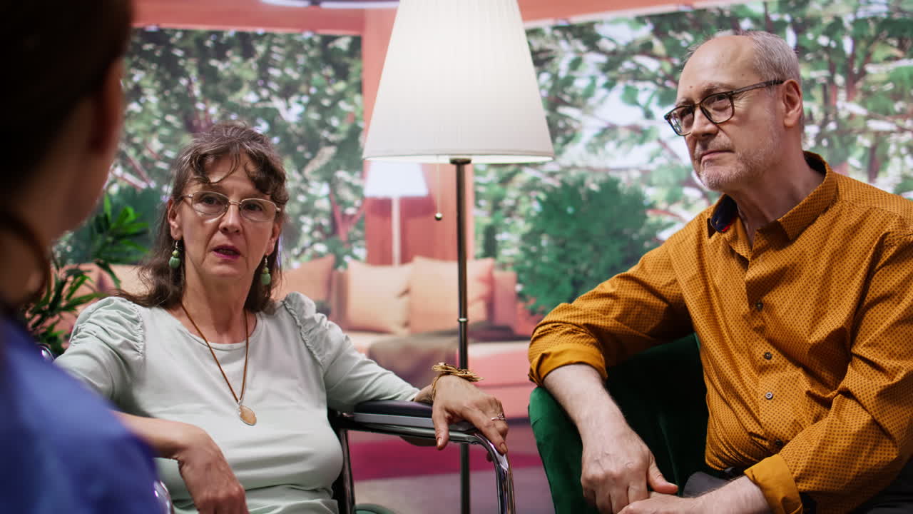 노인 은 간호사 와 대화 하는 동안 체어 에 앉아 장애 를 가진 아내 를 위로 한다