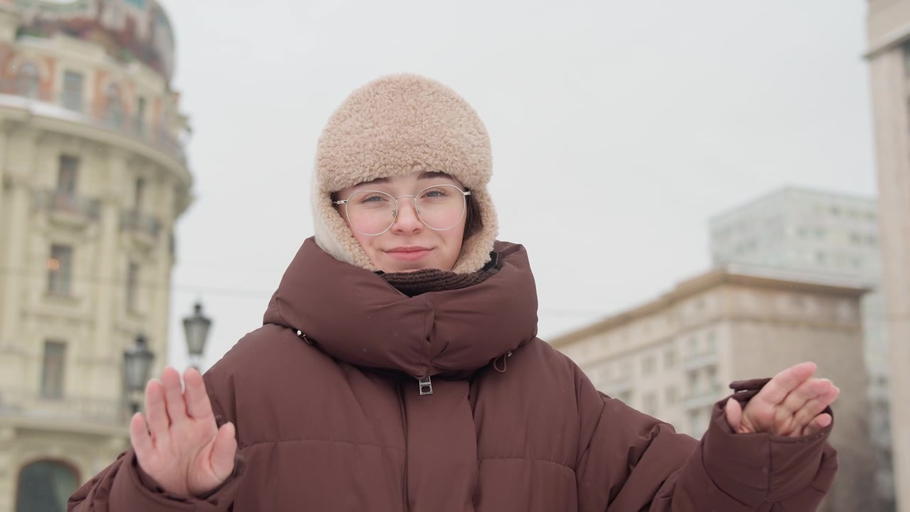 Nuori nainen ruskeassa pörröisessä talvitakissa ja beigessä turkislakissa seisoo lumisella kaupunkiaukiolla, pitäen käsilaukkua sivullaan, kädet kohotettuina rinnan lähellä ja katsoen oikealle rauhallisella ilmeellä pehmeän pilvisen valon alla.