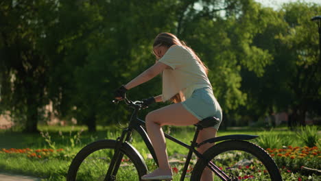 el ciclista se prepara para un paseo montando la bicicleta cerca de un jardín vibrante, posicionando la pierna izquierda en el pedal mientras comprueba la palanca de freno, el fondo muestra vegetación exuberante, flores coloridas y árboles altos