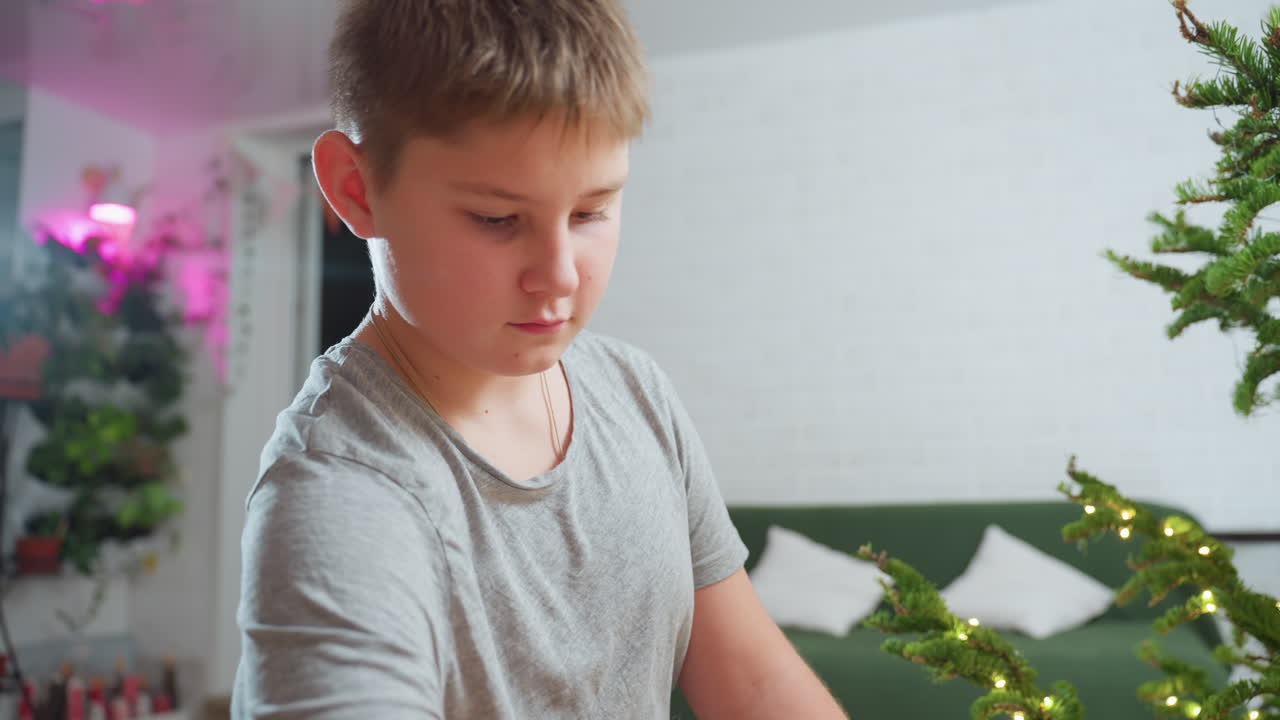 Boy in grey top standing beside Christmas tree decorated with fairy lights, carefully opening vintage box and smiling warmly as he brings out colorful ornament, cozy festive indoor scene