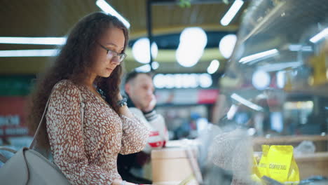 side view of woman in vintage dress standing in front of store holding menu paper, looking contemplatively at choices while another person beside her rests head on hand thoughtfully