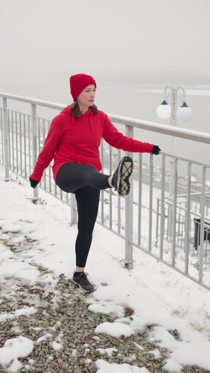woman exercising outdoors during winter holding iron railing for balance while stretching leg back and forth on snowy ground amid foggy atmosphere with distant river and light poles in background