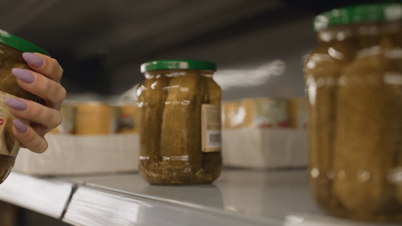Close up of manicured hand grasping glass jar of pickles from store shelf in bright grocery aisle highlighting green lid polished nails and retail selection with blurred background and metal rails