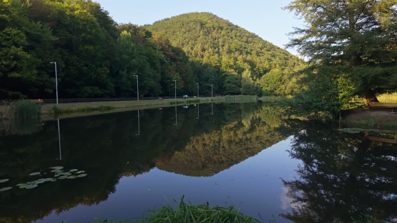 Tranquil view of Áfonyás-tó, reflecting the surrounding forested hills and a clear blue sky in Nagyhuta, Hungary, Zemplén