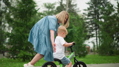 a caring mother is gently guiding her focused young son as he rides his bicycle along an interlocked pathway, she touches his head to offer support and help him maintain balance
