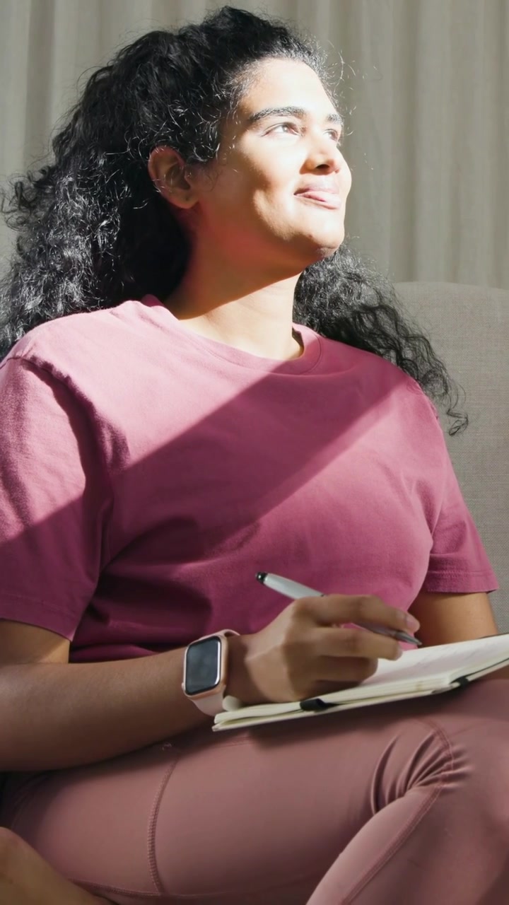 Video verticale: Donna che scrive, osserva il cambio di luce del sole sul divano, pensa con penna e blocco note, con maglietta bordeaux.