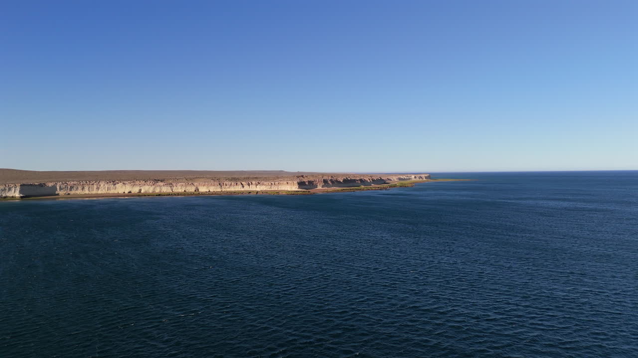 Experience a stunning aerial establishing shot over Puerto Madryn, Argentina, showcasing the vast Atlantic Ocean and picturesque coastline under a clear, bright blue sky.