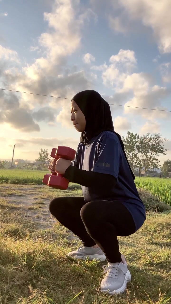 Video vertikal gerak lambat seorang wanita Muslim Asia berhijab sedang melakukan dumbbell goblet squat. Dia berolahraga di luar ruangan di lapangan hijau dengan latar belakang langit senja berawan yang indah