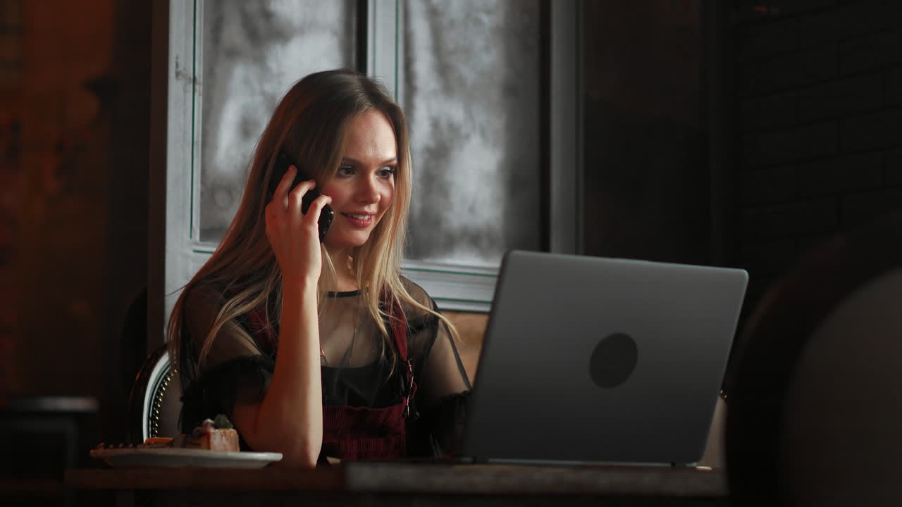 Ung kvinde sidder på café ved træbord, drikker kaffe og bruger smartphone. En bærbar computer står på bordet. Pige surfer på internettet, chatter og blogger. Kvinde holder telefon og kigger på skærmen.