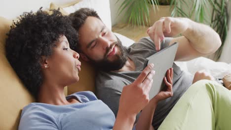 Video-De-Una-Feliz-Pareja-Diversa-Relajándose-En-Casa,-Acostada-En-La-Cama-Usando-Una-Tableta