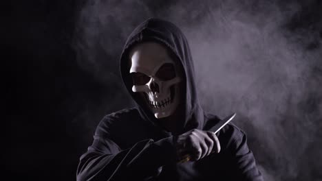 close up of scary man in the hooded sweatshirt wearing halloween mask holding a knife and making gesture want to kill someone crazy on the black background with smoke