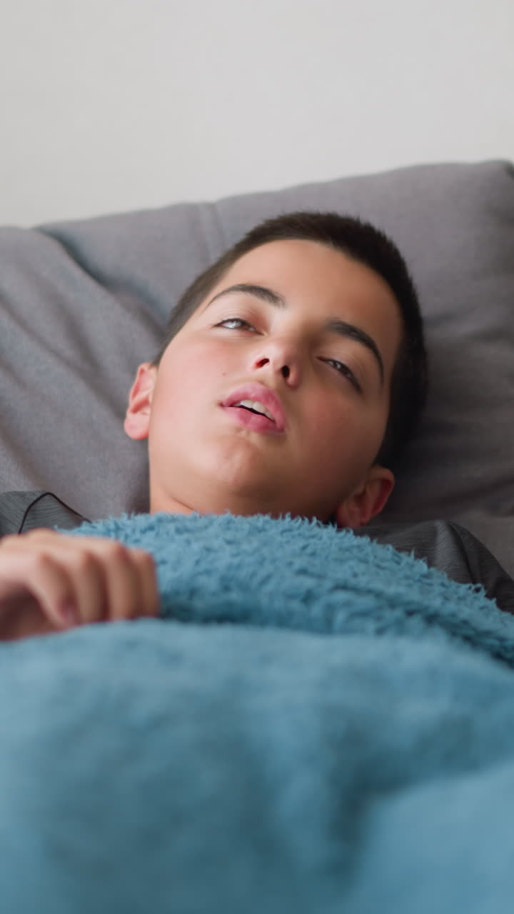 Sick student on holiday resting on couch wrapped in blanket adjusting for comfort with used tissue beside him, relaxing indoor setting with soft natural light streaming through window