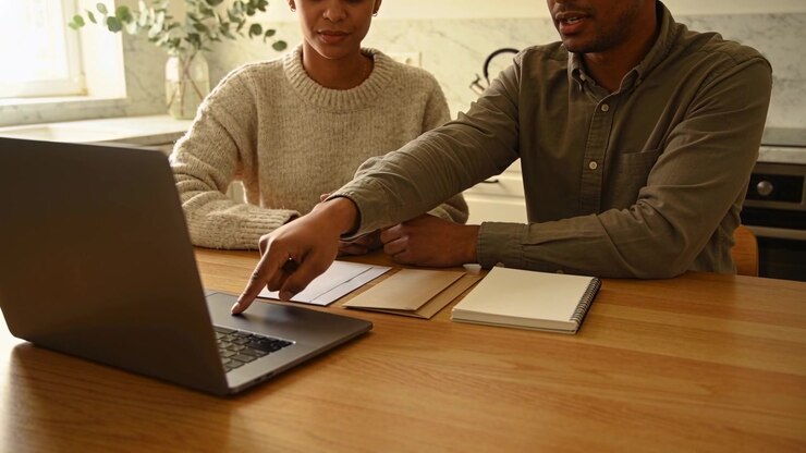 Woman and man sitting at wooden table, pointing at laptop screen then holding hands, AI-generated